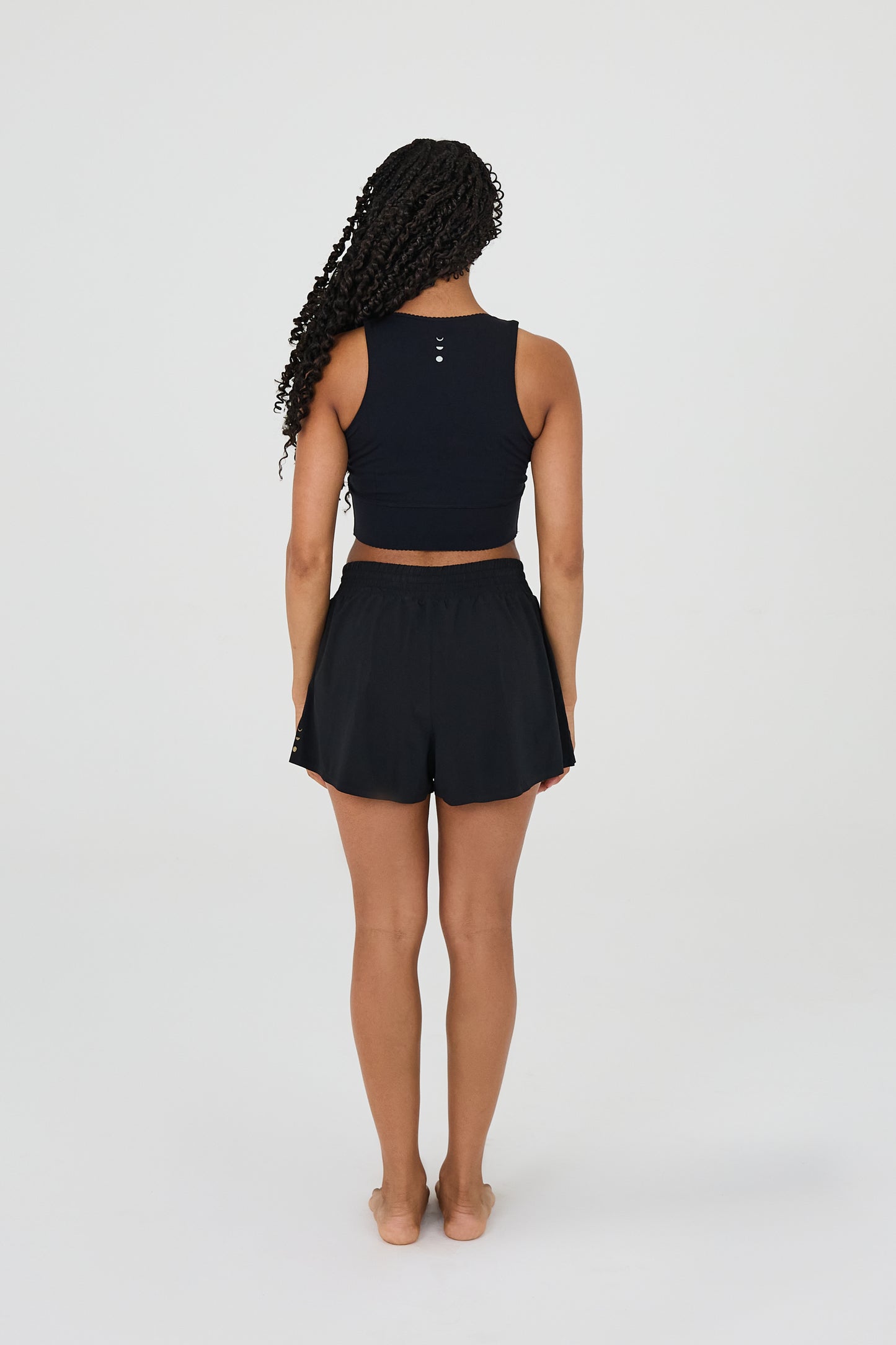 Vue de dos du modèle portant le short L’Original Flowy, composé d’une couche extérieure plissée ultra-légère et d’un short intérieur sculptant à compression. Ce design 2-en-1 allie fluidité féminine et maintien performant, avec l’intégration brevetée des MicroPerle® et une poche intérieure discrète pour l’essentiel, comme un téléphone ou une balle de tennis.