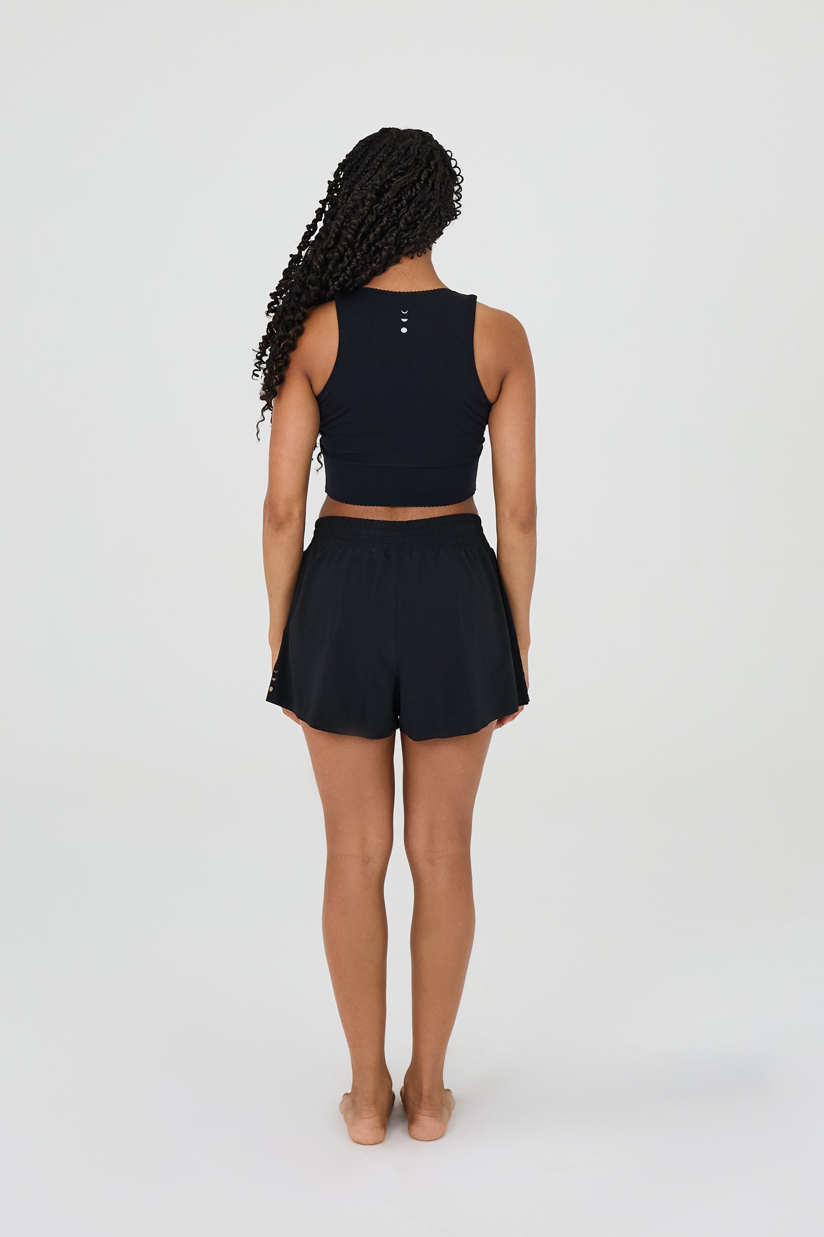 Vue de dos du modèle portant le short L’Original Flowy, composé d’une couche extérieure plissée ultra-légère et d’un short intérieur sculptant à compression. Ce design 2-en-1 allie fluidité féminine et maintien performant, avec l’intégration brevetée des MicroPerle® et une poche intérieure discrète pour l’essentiel, comme un téléphone ou une balle de tennis.