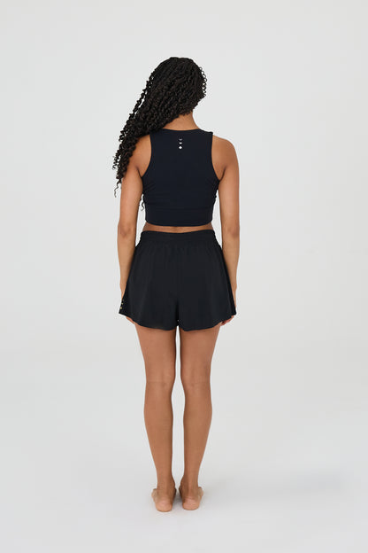 Vue de dos du modèle portant le short L’Original Flowy, composé d’une couche extérieure plissée ultra-légère et d’un short intérieur sculptant à compression. Ce design 2-en-1 allie fluidité féminine et maintien performant, avec l’intégration brevetée des MicroPerle® et une poche intérieure discrète pour l’essentiel, comme un téléphone ou une balle de tennis.