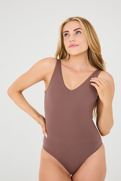 Modèle portant le body Adorn coloris cappuccino de la collection sculpturale Adorn d’Elastique, avec un décolleté flatteur orné de festons, un dos nu, et une silhouette lisse et ajustée, pensée pour être portée aussi bien en maillot qu’au quotidien. Confectionné dans un tissu UPF 50 et infusé de MicroPerle® brevetées pour une stimulation lymphatique douce et un maintien du centre du corps.