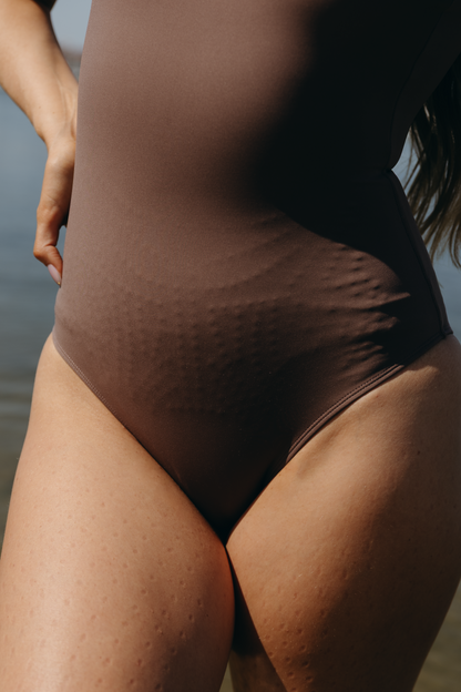 Modèle portant le body Adorn coloris cappuccino, issu de la collection sculpturale Adorn d’Elastique. Ce modèle présente un décolleté flatteur souligné d’une finition festonnée, un dos nu et une silhouette lisse et ajustée, conçue pour être portée aussi bien en maillot qu’au quotidien. Réalisé en tissu UPF 50 et infusé de MicroPerle® brevetées pour une stimulation lymphatique douce et un maintien du centre du corps.