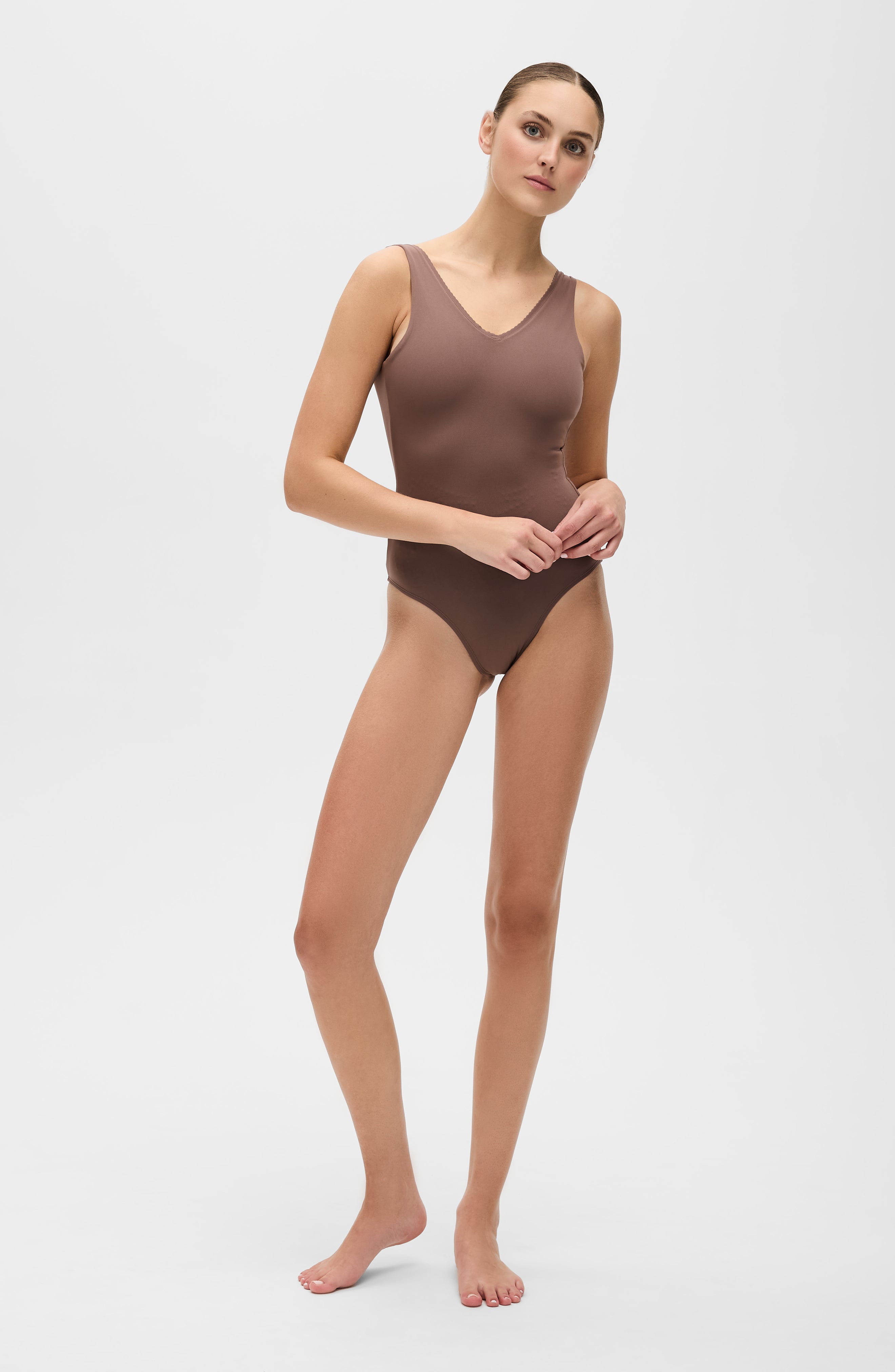 Modèle portant le body Adorn coloris cappuccino de la collection sculpturale Adorn d’Elastique, avec un décolleté flatteur orné de festons, un dos nu, et une silhouette lisse et ajustée, pensée pour être portée aussi bien en maillot qu’au quotidien. Confectionné dans un tissu UPF 50 et infusé de MicroPerle® brevetées pour une stimulation lymphatique douce et un maintien du centre du corps.