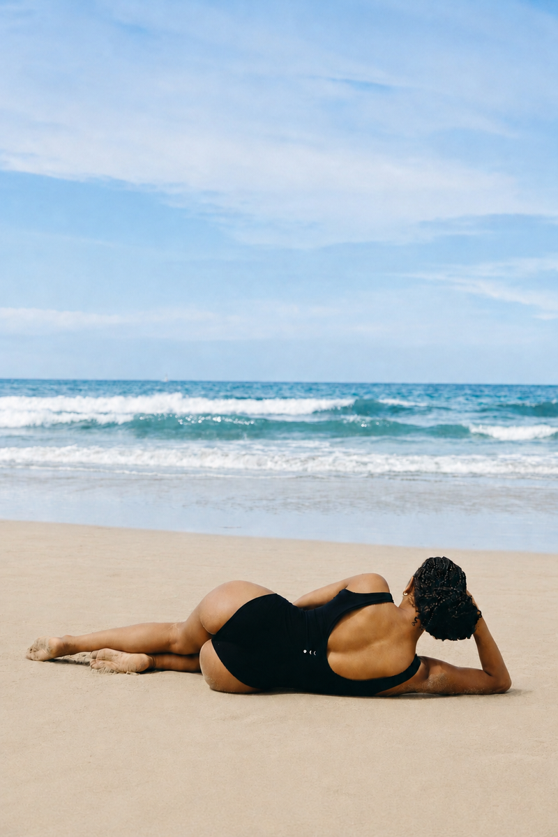 Modèle portant le body Adorn coloris noir, issu de la collection sculpturale Adorn d’Elastique. Il présente un décolleté flatteur bordé de festons, un dos nu et une silhouette lisse et ajustée, conçue pour être portée en maillot ou au quotidien. Confectionné dans un tissu UPF 50 et infusé de MicroPerle® brevetées pour une stimulation lymphatique douce et un maintien ciblé du centre du corps.