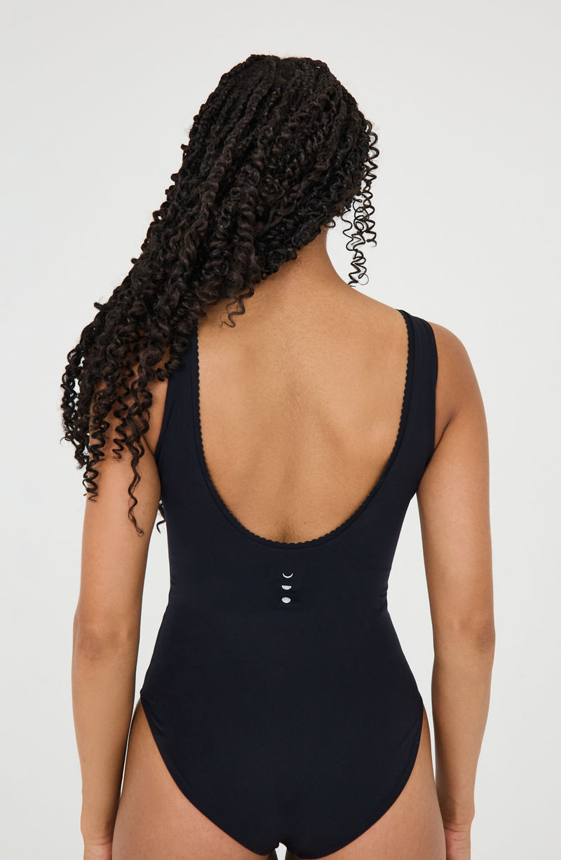 Modèle portant le body Adorn coloris noir, issu de la collection sculpturale Adorn d’Elastique. Il présente un décolleté flatteur bordé de festons, un dos nu et une silhouette lisse et ajustée, conçue pour être portée en maillot ou au quotidien. Confectionné dans un tissu UPF 50 et infusé de MicroPerle® brevetées pour une stimulation lymphatique douce et un maintien ciblé du centre du corps.