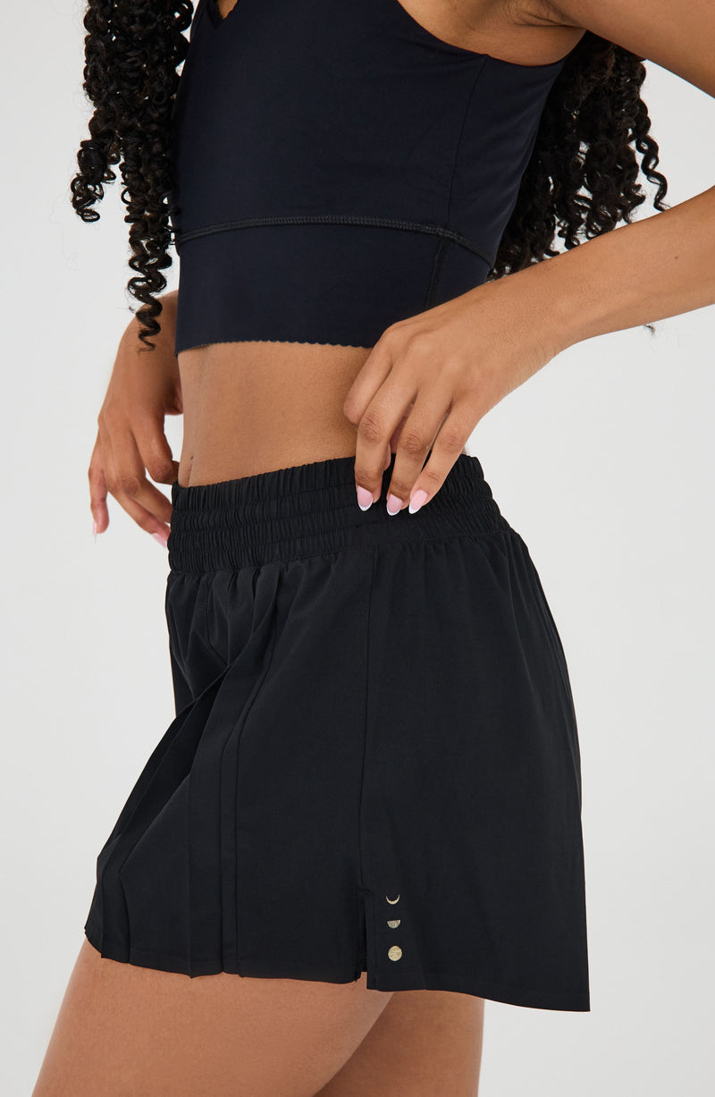 Modèle portant le short L’Original Flowy, composé d’une couche extérieure plissée ultra-légère et d’un short intérieur gainant à effet sculptant. Ce design 2-en-1 allie élégance féminine et maintien performant, avec l’intégration brevetée de MicroPerle® et une poche intérieure discrète pour y glisser l’essentiel — téléphone ou balle de tennis.