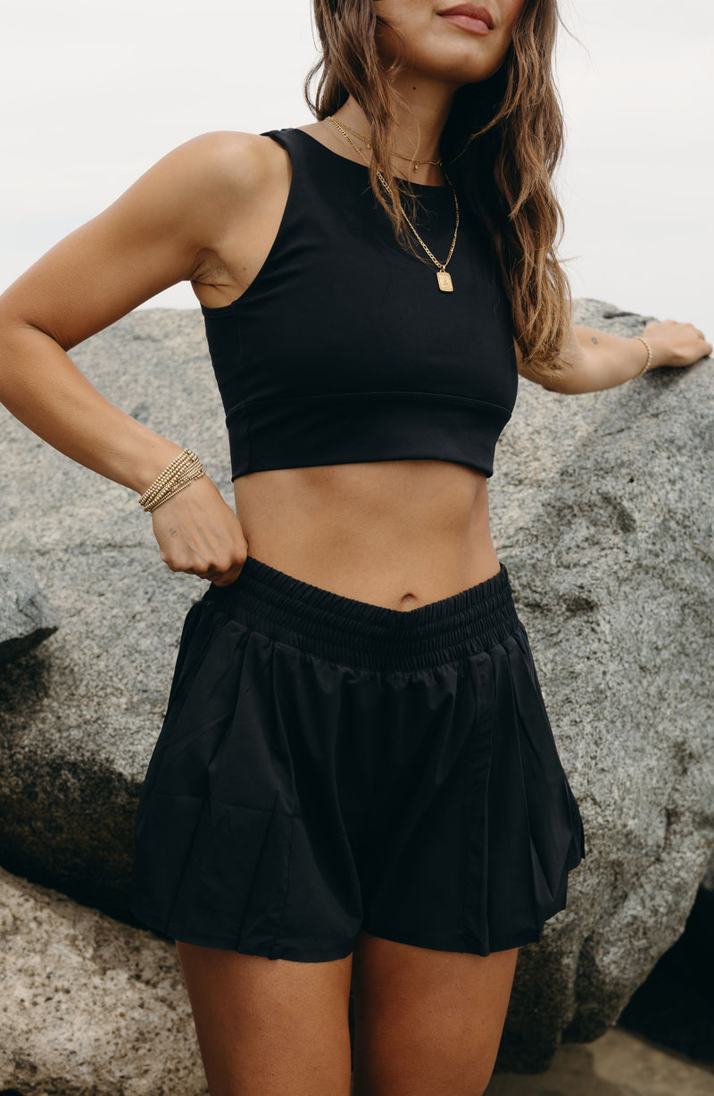 Modèle portant le short L’Original Flowy, composé d’une couche extérieure plissée ultra-légère et d’un short intérieur gainant à effet sculptant. Ce design 2-en-1 allie élégance féminine et maintien performant, avec l’intégration brevetée de MicroPerle® et une poche intérieure discrète pour y glisser l’essentiel — téléphone ou balle de tennis.