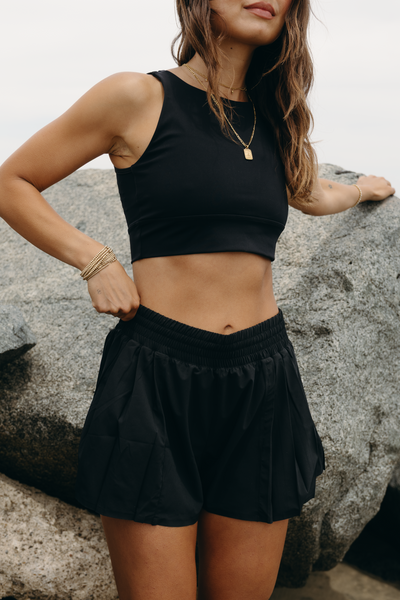 Modèle portant le short L’Original Flowy, composé d’une couche extérieure plissée ultra-légère et d’un short intérieur gainant à effet sculptant. Ce design 2-en-1 allie élégance féminine et maintien performant, avec l’intégration brevetée de MicroPerle® et une poche intérieure discrète pour y glisser l’essentiel — téléphone ou balle de tennis.
