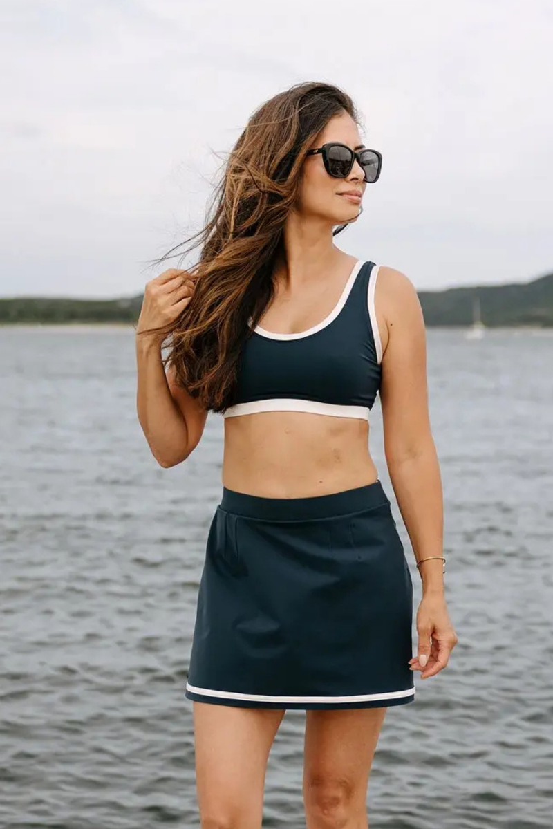 Femme portant le haut de bikini et la jupe-short compressifs bleu marine et crème de la collection Monte Carlo avec technologie MicroPerle® intégrée. Conçu pour favoriser le drainage lymphatique, offrir une compression sculptante et allier confort et élégance, cet ensemble inspiré de l’univers balnéaire haut de gamme accompagne naturellement les mouvements au bord de l’eau.